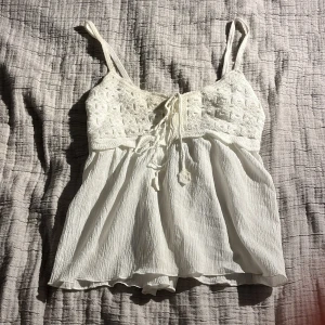 Vit ärmlös topp med spetsdetaljer - Säljer en söt vit ärmlös topp med spetsdetaljer och knytning framtill. Toppen har tunna axelband och ett luftigt fall, perfekt för varma dagar. Den har en smockad rygg för extra komfort och stil.