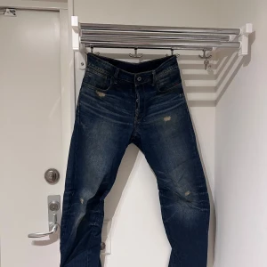 Mörkblå jeans med slitningar Gstar - Snygga mörkblå jeans med coola slitningar på framsidan. De har en klassisk femficksdesign och knappgylf. Perfekta för en avslappnad stil.