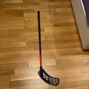 Innebandyklubba Salming med blått blad - Säljer en innebandyklubba Salming med ett blått blad och skaft i blått och rött. Perfekt för innebandyspelare som vill ha en pålitlig klubba med bra grepp och kontroll. Bladet har ett rutmönster för bättre bollhantering.