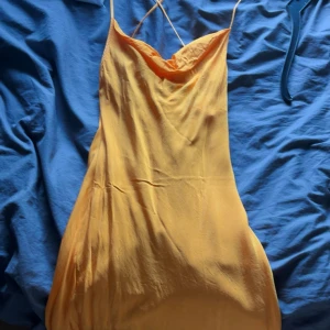 Orange klänning i satin från H&M - Snygg orange klänning från H&M i satinliknande material. Klänningen har tunna axelband, cowlneck-ringning och en figurnära passform. Perfekt för dig som vill sticka ut med en färgstark look och älskar trendiga siluetter. Banden korsar i ryggen. Tredje bilden är lånad för att visa hur den sitter vid bröten då jag sjöng inte kan visa den på då den är för liten. 