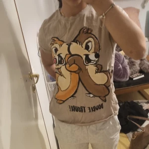 Beige Disney t-shirt med Chip & Dale - Säljer en beige t-shirt med stort tryck av Chip & Dale och texten 'Double Trouble' framtill. T-shirten är kortärmad och har en avslappnad passform. Perfekt för dig som älskar klassiska Disney-karaktärer och vill ha en rolig och unik look.