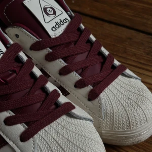 Adidas Superstar vit & vinröd sneaker - Adidas Superstar sneakers i vitt skinn med vinröda detaljer, klassiska tre ränder och snörning. Ikonisk gummitå och Adidas-logga på plös och häl. Perfekt för dig som vill ha en tidlös och snygg sneaker med streetkänsla.