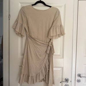 Beige omlottklänning från NA-KD - Supersöt beige kortklänning från NA-KD x Queen of Jetlags med volangdetaljer på ärmar och nederkant. Omlottmodell med knyt i midjan och korta, vida ärmar. Perfekt för en somrig look och till sneakers eller sandaler.