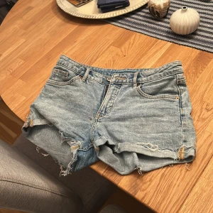 Ljusblå jeansshorts med fransar - Säljer ett par ljusblå jeansshorts med råa, fransiga kanter och slitna detaljer. Klassisk femficksmodell med bälteshällor och dragkedja. Perfekta för varma dagar när du vill ha en avslappnad och cool stil.