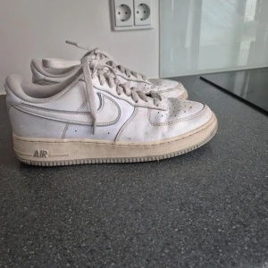 Vita Nike Air Force 1 sneakers - Klassiska vita Nike Air Force 1 sneakers med lågt skaft och snörning. Skorna har en platt sula, rund tå och är tillverkade i skinn. Perfekta för dig som gillar en clean och tidlös streetstil.