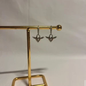 Silverörhängen med hjärtan och vingar - Snygga örhängen i silverfärg med hängande hjärtan som har små vingar och ett nyckelhål i mitten. Perfekta för dig som gillar unika detaljer och vill sticka ut lite extra. Passar till alla tillfällen och ger en cool vibe till din stil.