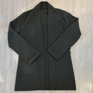 Grön kavaj från KDN Moda, stl S - Stilren tunn grön kavaj från KDN Moda i storlek S. Jackan har en rak och enkel design utan knappar, med långa ärmar och mjukt tyg som ger en clean look. 