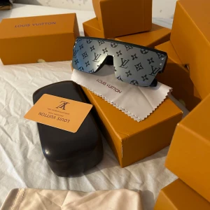 Lv glasögon  - Snygga och futuristiska solglasögon. design med svarta bågar och ikoniska LV-detaljer. Perfekt för dig som vill sticka ut och köra en lyxig vibe.