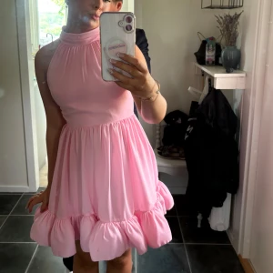 Rosa kort klänning med volang från MALINA - Supersöt rosa kortklänning med hög hals och knytband i nacken. Klänningen har en volangkant nertill och är ärmlös, så himla fin älskar den verkligen🩷 andvänd 1 gång, ny pris cirka 2599! Den är storlek xs men passar mig som har S/M