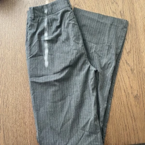 Grå randiga kostymbyxor Madlady 36 Tall - Snygga grå kostymbyxor från Madlady i storlek 36 Tall. Byxorna har tunna vita ränder, klassisk passform med hällor för bälte och stängs med dragkedja och hyska. Midwaist