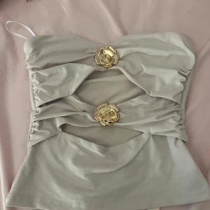 Beige tubtopp med gulddetaljer - Superfin beige tubtopp med två stora guldfärgade blomdetaljer framtill. Toppen har snygga draperingar och cut-outs som ger en unik look. Perfekt för dig som vill sticka ut med en trendig och elegant topp.