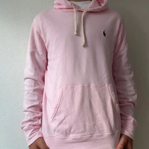 Tunn Ljusrosa hoodie från Polo Ralph Lauren - Säljer en ljusrosa hoodie från Polo Ralph Lauren med klassisk broderad logga på bröstet. Tröjan har huva med snörning, känguruficka och är tillverkad i mjuk bomull. Perfekt för dig som gillar stilren och chill streetwear. Modellen på bilden är 174cm och väger ca 65kg.