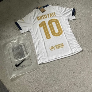 FC Barcelona Ansu Fati vit matchtröja Nike - Snygg FC Barcelona fotbollströja i vitt med guldiga detaljer och tryck, Ansu Fati #10 på ryggen. Tröjan har klubbmärke, Nike-logga och UNHCR-tryck. Mörkblå krage och ärmslut. Tillverkad i lätt och ventilerande material, perfekt för match eller träning.