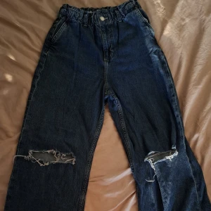 Wide leg jeans med hål &Denim - Mörkblå wide leg jeans från &Denim med hög midja och resår i midjan. Snygga slitningar och hål på båda knäna ger en cool, avslappnad vibe. Perfekta för dig som gillar en loose och trendig passform.