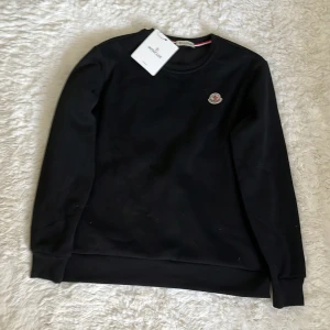 Svart Moncler sweatshirt i bomull - Svart sweatshirt från Moncler med klassisk rund halsringning och Moncler-logga på bröstet. Tröjan har långa ärmar, ribbade muddar och är tillverkad i mjuk bomull. Perfekt för en clean och stilren look.