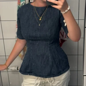 Mörkblå jeansblus med figursydd passform - Supersnygg mörkblå jeansblus med kort ärm och rund hals. Figursydd modell som ger en markerad midja och cool siluett. Perfekt att styla med både kjol och byxor för en trendig look. Materialet är klassiskt jeans och toppen har en enkel, stilren design.