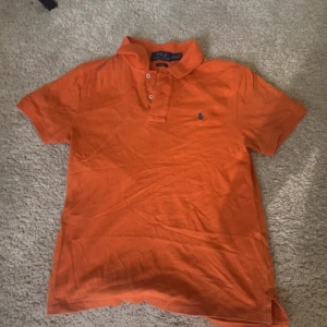 Orange pikétröja Polo Ralph Lauren - Snygg orange pikétröja från Polo Ralph Lauren i slim fit-modell. Storlek 14-16 sitter som XS. Broderad blå logga på bröstet. Tillverkad i mjuk bomull, perfekt för en fräsch och sportig look. 