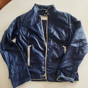 Blå vindjacka från North Sails L - Snygg marinblå vindjacka från North Sails med vita detaljer och dragkedjor. Jackan har hög krage med tryckknappar, två fickor med dragkedja och vita kanter längs ärmar och nederkant. Perfekt för blåsiga dagar och stilren look.