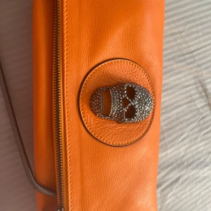  Thomas Wylde - Cool orange kuvertväska i skinn från Thomas Wylde med en blingad dödskalle-detalj framtill och metallplatta med logga på baksidan. Väskan har dragkedja och en edgy vibe som sticker ut i mängden. Nypris 8000 mitt pris 1700 billigare vid snabb affär