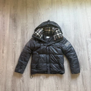 Svart pufferjacka från Burberry - Svart pufferjacka från Burberry med klassiskt rutigt foder i huvan. Jackan har dragkedja, knappar och två fickor framtill. På ärmen finns en patch med Burberry-logga. Perfekt för vintern/hösten och riktigt snygg.