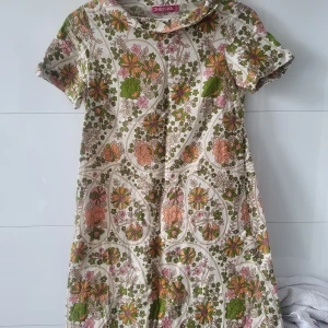 Blommig kortklänning från Indiska - Retroinspirerad kortklänning från Indiska med färgglatt blommönster i grönt, orange, rosa och beige. Klänningen har korta ärmar, lätt utställd passform och en liten krage. Perfekt för dig som gillar vintage vibes och unika prints.