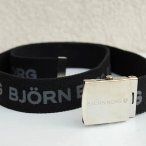 Svart tygbälte från Björn Borg - Svart tygbälte från Björn Borg med grå logotyp tryckt längs hela bandet. Bältet har en stilren, silverfärgad metallspänne med ingraverad logga. Perfekt accessoar för en sportig och avslappnad look.