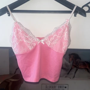 Rosa linnetopp med spetsdetaljer - Supersöt rosa linnetopp med tunna axelband och vita spetsdetaljer upptill. Toppen har en liten rosett framtill och är croppad i modellen. Perfekt för dig som gillar en feminin och trendig stil. Säljer billigt, har ingen användning av den💓