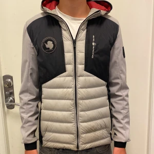 Sail Racing grå och svart pufferjacka - Snygg pufferjacka från Sail Racing i grått och svart med röd insida. Jackan har quiltad framsida, dragkedja, luva och coola logodetaljer på bröstet. Perfekt för dig som vill ha en sportig och modern look.