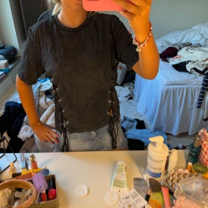 Svart t-shirt med snörning i sidorna - Cool svart t-shirt med tvättad look och snörningsdetaljer längs båda sidorna. T-shirten har korta ärmar och en avslappnad passform, perfekt till jeans eller shorts för en edgy vibe.