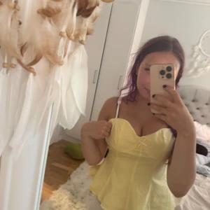 Gul korsett-topp med rosett - Söt gul korsett-topp med liten vit rosett framtill och volangig nederkant. Axelbandslös design med markerad byst och lätt utsvängd passform. Perfekt för dig som vill sticka ut med en färgglad och feminin look.