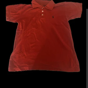 Röd pikétröja från Polo Ralph Lauren - Klassisk röd pikétröja från Polo Ralph Lauren med broderad logga på bröstet. Tröjan har krage, tre knappar framtill och korta ärmar. Tillverkad i mjuk bomull som är skön mot huden. Perfekt för en avslappnad och stilren look.
