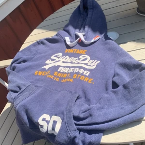 Blå Superdry hoodie med tryck - Blå hoodie från Superdry med stort vitt och gult tryck på bröstet och siffran 60 på ärmen. Klassisk huva med snörning och känguruficka framtill. Mjuk bomullsblandning, perfekt för chill dagar. Snygg streetstyle-vibe!
