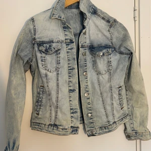 Ljusblå jeansjacka med silvriga knappar - Snygg ljusblå jeansjacka med klassisk tvätt och silvriga knappar framtill. Jackan har två bröstfickor med lock och knapp, samt två sidofickor. Perfekt för lager-på-lager och enkel att styla till olika outfits.