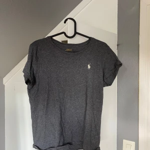 Mörkgrå t-shirt från Polo Ralph Lauren - Snygg mörkgrå t-shirt från Polo Ralph Lauren med klassisk vit logga broderad på bröstet. T-shirten är i mjuk bomull och har rund halsringning och korta ärmar. Perfekt basic-plagg med stilren look.