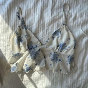 Blommig volang-linnetopp i blått och vitt - Supersöt linnetopp med volangkant och smala axelband. Toppen är vit med blått blommönster och har en lätt och luftig känsla, perfekt för varma dagar. V-ringad framtill och med justerbara band för bästa passform. Ger en romantisk och somrig vibe.