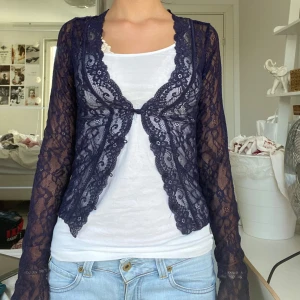 Mörkblå spetskofta med lång ärm - Säljer en mörkblå, transparent spetskofta med långärmad design. Koftan har vacker spets med blommönster och en öppen front som knyts framtill. Perfekt att slänga över ett linne eller t-shirt för en extra touch till outfiten.