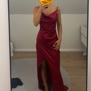 Vinröd långklänning med slits - Elegant vinröd långklänning i satinliknande material med smala axelband och draperad överdel. Klänningen har en snygg slits framtill och figurnära passform som framhäver siluetten. Perfekt för bal eller festliga tillfällen.