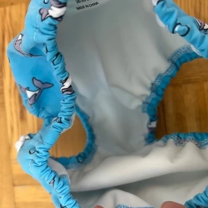 Blå bikiniunderdel med delfinmotiv - Söt blå bikiniunderdel från Dolphin Swimwear med gulligt delfinmönster och små bubblor. Modellen har resår i midja och ben, samt praktiska knappar i sidorna för justering. Perfekt för strandhäng och bad under sommaren!