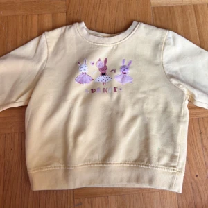 Gul sweatshirt med dansande kaniner - Supersöt ljusgul sweatshirt med tryck av tre dansande kaniner i lila och rosa kjolar framtill. Tröjan har rund hals, långa ärmar och mjuk mudd vid ärmslut och nederkant. Perfekt för dig som gillar gulliga motiv och mjuka material.
