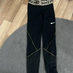 Nike Pro svarta tights med guldiga detaljer - Nike Pro tights i svart med bred beige resår i midjan och svart Nike-logga. Snygga guldiga sömmar och meshdetaljer nedtill på benen. Perfekta för träning och riktigt sköna att ha på sig. Tight passform och stretchigt material. Det står mitt namn på lappen det är det enda som är ovant med byxorna.