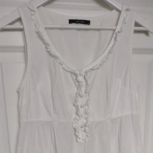 Vit/benvit Y2K topp/tunika från Vero Moda - Superfin benvit lite längre topp från Vero Moda med volangdetaljer och knappar framtill. Ärmlös modell i tunn, randig bomull med romantiska volanger vid halsringning och nederkant.  Storlek S. Väldigt snygg till jeansshorts/kjol, linnebyxor m.m.