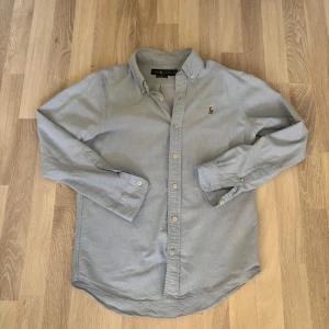 Ljusblå skjorta från Ralph Lauren S - Klassisk ljusblå skjorta från Ralph Lauren i bomull med broderad logga på bröstet. Skjortan har knappar hela vägen, button-down-krage och långa ärmar. Custom fit-modell som sitter snyggt och passar till många olika stilar.