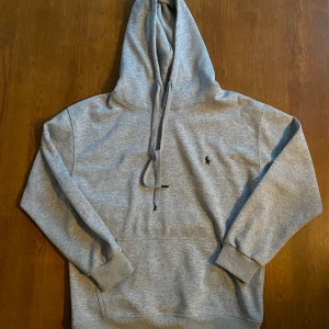 Grå hoodie från Polo Ralph Lauren - Snygg grå hoodie från Polo Ralph Lauren med klassisk broderad logga på bröstet. Tröjan har huva med snörning, känguruficka och ribbade muddar. Perfekt för chill dagar eller när du vill ha en clean och stilren look. Står M men sitter mer som typ S