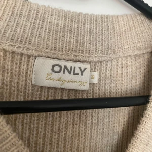 Beige stickad väst från ONLY - Säljer en beige stickad väst från ONLY med bred ribbad kant nedtill och vid ärmöppning. Västen har en rund halsringning och är i en mjuk, grovstickad kvalitet som är perfekt för lager-på-lager-stil.