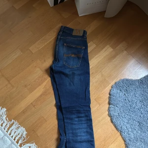 Mörkblå jeans från Nudie Jeans - Säljer ett par mörkblå jeans från Nudie Jeans med klassisk femficksdesign och snygga slitningar. Jeansen har orange sömmar och Nudie-logga på bakfickan. Modellen är slim och sitter snyggt längs benen. Perfekta för dig som gillar stilrena och bekväma jeans.