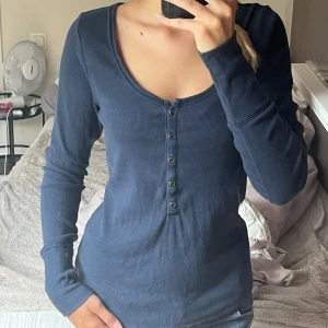 Mörkblå ribbad tröja Hollister L - Superskön mörkblå långärmad tröja från Hollister i ribbad bomull. Djup rundringning och snygga knappar framtill. 