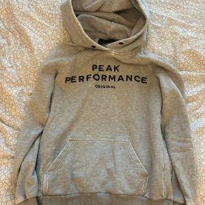 Grå hoodie från Peak Performance XS - Snygg ljusgrå hoodie från Peak Performance i storlek XS. Tröjan har svart broderad logga på bröstet, stor huva och en klassisk känguruficka framtill. Perfekt för chill dagar eller när du vill ha en sportig look.