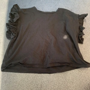 Svart volangärmad topp från Zara - Svart topp från Zara i storlek S med korta ärmar och snygga volangdetaljer på axlarna. Tillverkad i mjuk bomull som känns skön mot huden. Perfekt för dig som gillar en enkel men ändå trendig look.