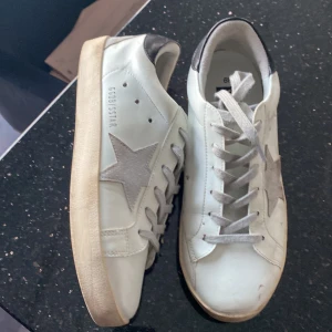 Vita Golden Goose sneakers med stjärna - Golden Goose sneakers i vitt skinn med grå mockastjärna på sidan och svarta detaljer vid hälen. Klassisk rund tå, platt sula och grå snörning. Snygg och avslappnad look med ikonisk stjärndetalj på sidan. Pris kan diskuteras vid snabb affär