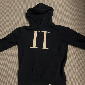Marin blå hoodie från Les Deux med tryck - Marin blå hoodie från Les Deux med stor vit II-logga på bröstet. Klassisk modell med huva och långa ärmar, ribbade muddar och mjukt material. Perfekt för dig som gillar stilrena och enkla plagg med en twist. Två små skador på höger ärm som visas på bild 4 och 5.